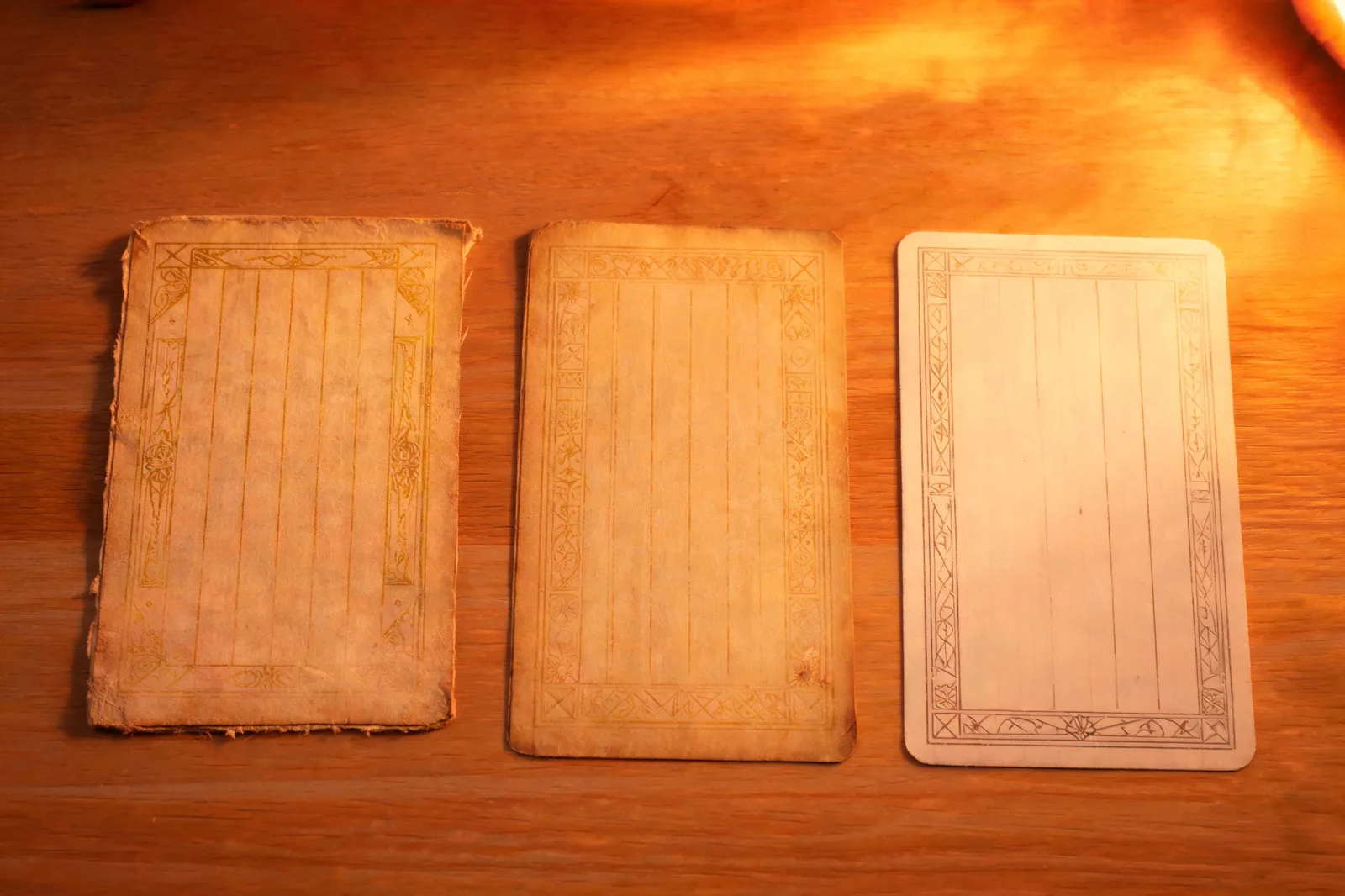 Three worn tarot papers laid side by side on a plain wooden desk in warm firelight, an early 1900s journal page, a mid-century book page and a modern card, book t golden dawn printings